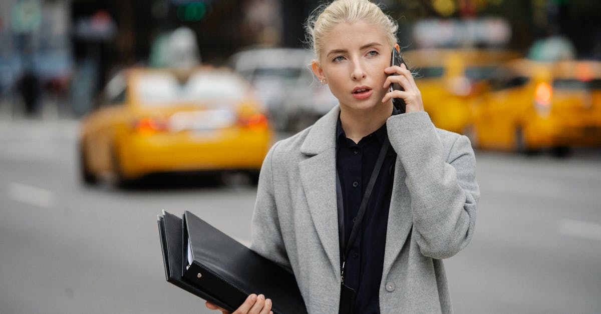Focused young businesswoman in gray coat standing on busy street with folders and having conversation on mobile phone
